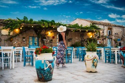 Young Woman Dressed In Romantic Dress And Sun Hat Walks Near The Colorful Outdoor Cafe At Main Square In The Beautiful Sicilian Coastal Village Marzamemi In Sicily At Sunrise With Dramatic Sky