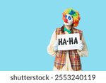 Young woman dressed as sad clown holding card with text HA-HA on blue background. April Fool