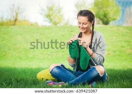 A young woman dressed in trendy jeans, a cardigan and with a backpack sitting in a city park on green grass and knitting a green sweater with knitting needles on a summer day