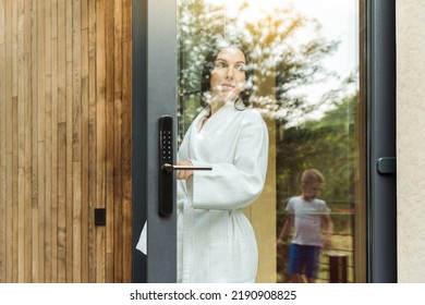 Young Woman Dressed In White Bathrobe Leaving The Modern House On The Terrace, Looking After Her Child Through The Glass Of The Door, Enjoying Beautiful Outdoors Atmosphere.