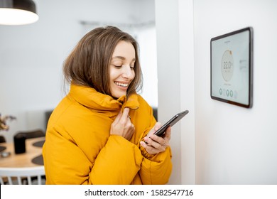 Young woman dressed in winter jacket feeling cold at home, controlling heating temperature with a digital tablet of a smart home