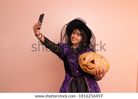A young woman dressed in a witch costume posing and photographing a selfie with a plain background