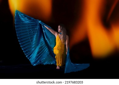 
Young Woman, Dressed In Yellow With Blue Isis Wings, With A Background Of Fire