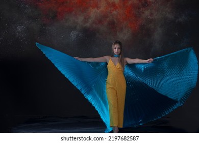 

Young Woman, Dressed In Yellow Dancing With Blue Isis Wings, With A Background Of Fire