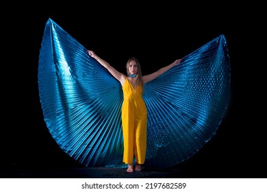 

Young Woman, Dressed In Yellow Dancing With Blue Isis Wings, With A Background Of Fire