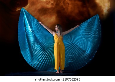 

Young Woman, Dressed In Yellow Dancing With Blue Isis Wings, With A Background Of Fire