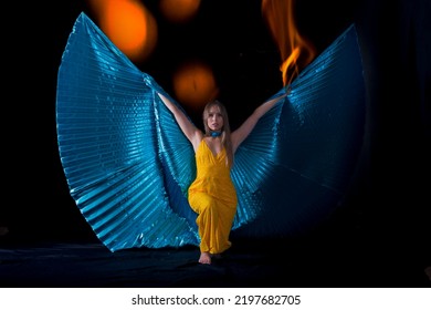 
Young Woman, Dressed In Yellow Dancing With Blue Isis Wings, With A Background Of Fire