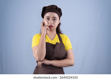 A young woman dressed in a yellow shirt and brown apron looks thoughtful and slightly sad, resting her hand on her cheek while standing against a plain background. - Powered by Shutterstock - Get 15% off with code: PIKWIZARD15