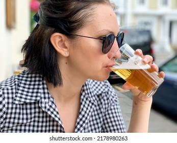 Young Woman Is Drinking Beer In A Cafe Or Restaurant Pub Outdoor. Drinking Alcohol. Candid. Refreshment, Summer Time, Pub Garden. Wearing Black And White Checkered Dress And Sunglasses. Drinking Lager