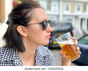 Young Woman Is Drinking Beer In A Cafe Or Restaurant Pub Outdoor. Drinking Alcohol. Candid. Refreshment, Summer Time, Pub Garden. Wearing Black And White Checkered Dress And Sunglasses. Drinking Lager