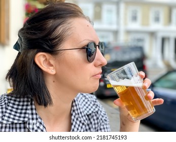 Young Woman Is Drinking Beer In A Cafe Or Restaurant Pub Outdoor. Drinking Alcohol. Candid. Refreshment, Summer Time, Pub Garden. Wearing Black And White Checkered Dress And Sunglasses. Drinking Lager