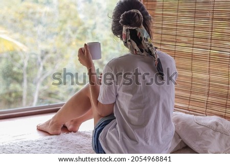 young woman drinking coffee and admiring the forest view in a wooden cabin - exotic lodging experience.
