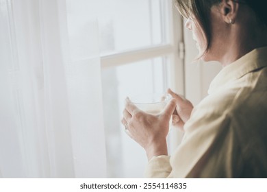 Young woman drinking a cup of tea looking outside the window Portrait of a girl enjoying free time at home. Middle aged female with a drink in the hand look through the door. Lifestyle leisure concept - Powered by Shutterstock