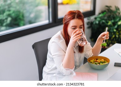Young woman drinking a glass of water while eating a salad at work. - Shutterstock ID 2134323781