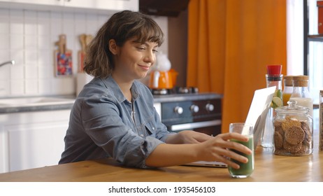 Young woman drinking green juice detox cocktail in the kitchen at home and working on the computer. Working from home and living healthy. - Powered by Shutterstock