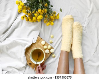 Young Woman Drinking Herbal Tea With Yellow Candies Wearing Yellow Cozy Socks In Cozy Bed With Yellow Spring Flowers In The Morning Or At Holidays