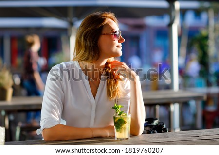 Young woman drinking mojito cocktail at cafe terrace at hot summer day. Beautiful businesswoman enjoying warm evening at afterwork restaurant. Happy smiling alone lady.