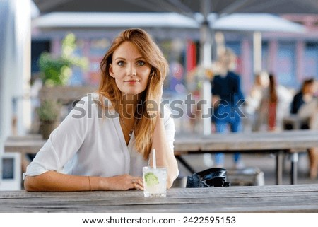 Young woman drinking mojito cocktail at cafe terrace at hot summer day. Beautiful businesswoman enjoying warm evening at afterwork restaurant. Happy smiling alone lady.