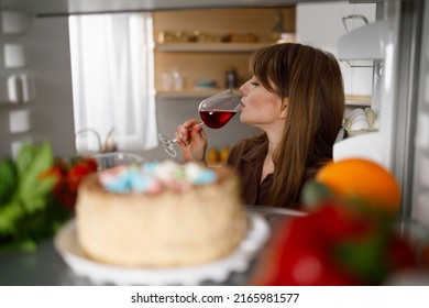 Young Woman Drinking Red Wine Standing By The Open Fridge At The Kitchen