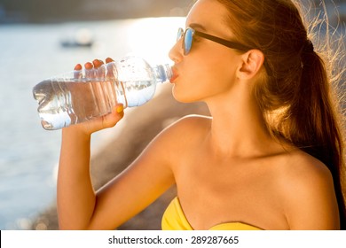 Young Woman Drinking Sparkling Water From Transparent Bottle On The Beach On Sunset