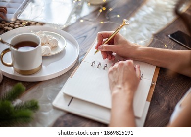 Young Woman Drinking Tea And Writing Plans Or Goals For New Year 2021 While Her Daughter Crafting Christmas Trees From Paper Cone, Yarns And Buttons With Placed Stars And Fairy Lights On Wooden Table.