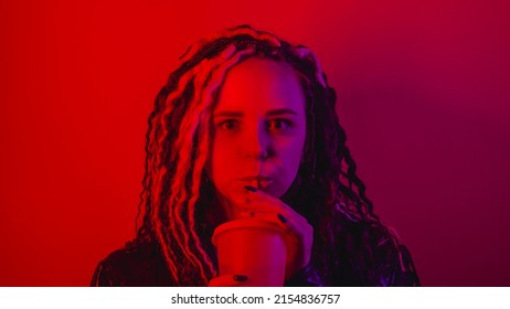 Young woman drinking through soft drink straw from large cup in dark room. Portrait of female enjoying nonalcoholic beverage in darkness illumination of red light - Shutterstock ID 2154836757