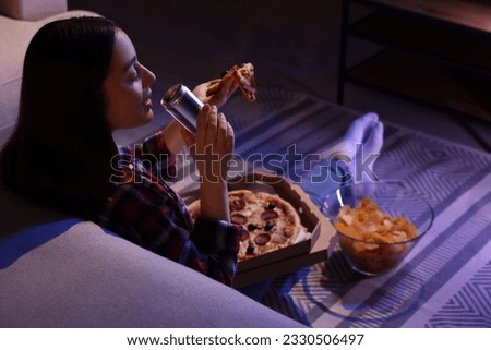 Young woman drinking from tin can and eating pizza while watching TV in room at night. Bad habit