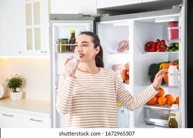 Young Woman Drinking Water Near Open Refrigerator In Kitchen