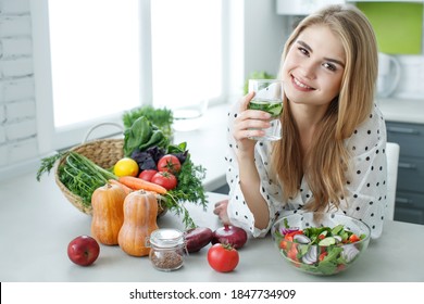 Young woman drinking water near the table with fruits and vegetables in the kitchen. Healthy food, drinks, diet, detox and people concept. High quality photo.