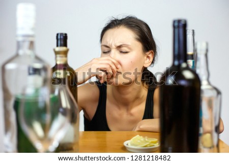 A young woman drinks alcohol at a table among the empty bottles of a room at home. Alcohol addiction makes her miserable. Portrait, background.