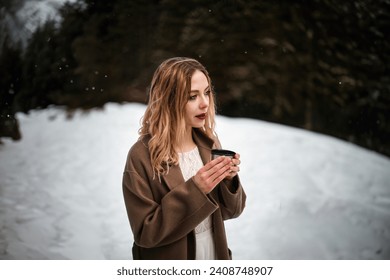 A Young woman drinks a hot beverage in the winter forest. Beautiful Girl heating her hands with tea or coffee cup during snowfall. Enjoying a warming drink in nature. - Powered by Shutterstock