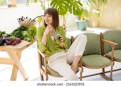 Young woman drinks lemonade while sitting on chair near table with lots of fresh food ingredients in room with green plants. Healthy lifestyle concept - Shutterstock ID 2161023935