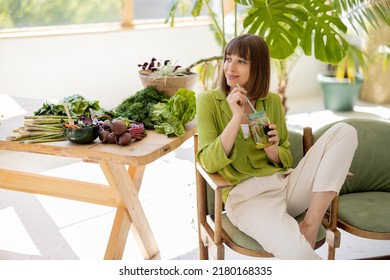 Young woman drinks lemonade while sitting on chair near table with lots of fresh food ingredients in room with green plants. Healthy lifestyle concept - Shutterstock ID 2180168335