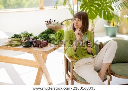 Young woman drinks lemonade while sitting on chair near table with lots of fresh food ingredients in room with green plants. Healthy lifestyle concept