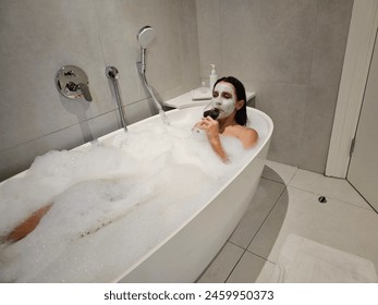 Young woman drinks red wine in a bubble bath. Facial rejuvenation mask and relax after a hard day - Powered by Shutterstock