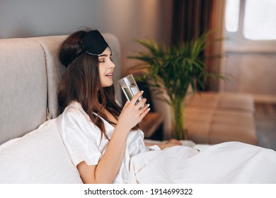 Young Woman Drinks Water In The Bed. Model With Brown Hair With A Glass Of Water In The Bed In The Bedroom. Drink A Glass Of Water In The Morning Or Before Bed. Young Woman Enjoying Drinking Water