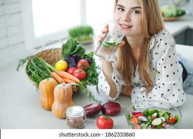  A young woman drinks water near the table with fruits and vegetables in the kitchen. High quality photo. - Shutterstock ID 1862333860