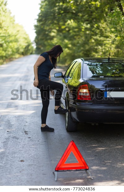 Young woman driver checking stopped\
car\'s wheels flagged by a red reflective\
triangle.