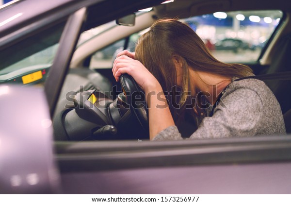 Young woman driver\
holding her head on the steering wheel in the car while sitting on\
the driver\'s seat maybe crying or feeling sad or angry on the\
parking in the night