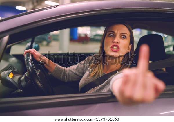Young woman driver showing middle finger trough\
the window of the car mad angry furious pissed on the parking in\
the city at night driving
