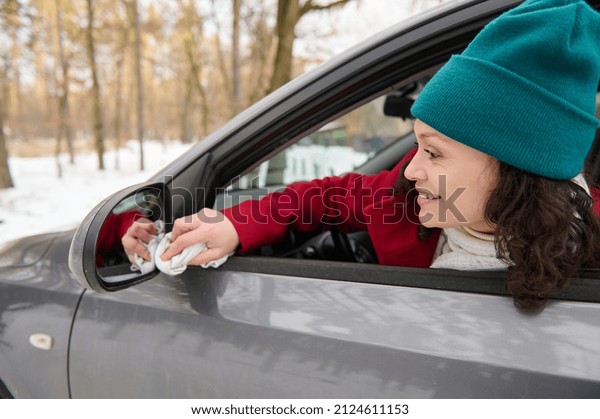 A young woman driver sits in the driver\'s seat of her\
car and wipes the driver\'s side mirror while stopping in a\
snow-covered forest landing while traveling on a beautiful cold\
snowy winter day