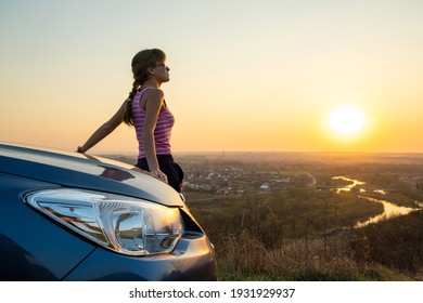 Young woman driver standing near her car enjoying warm sunset view. Girl traveler leaning on vehicle hood looking at evening horizon.