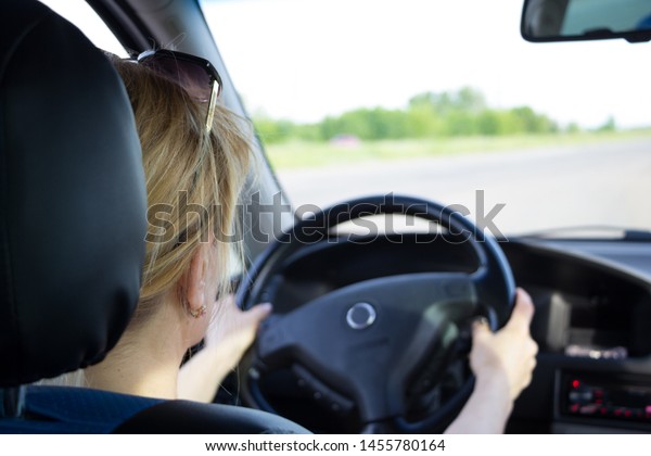 Young woman driving car on a highway. View from the\
back. Both hands on the steering wheel. Selective focus on her head\
with sun glasses on it. Safe driving concept. Black car interior.\
Road in blur. 