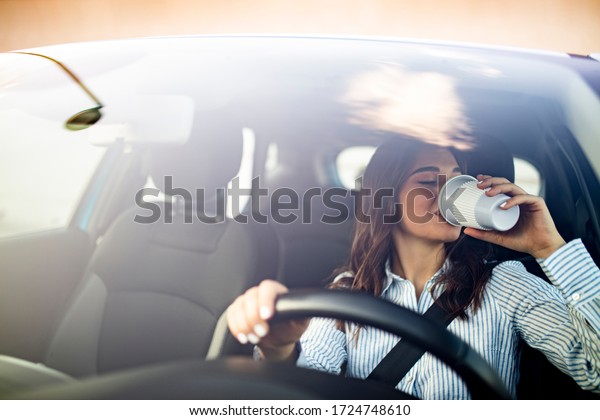 Young woman driving car with\
safety belt and drinking coffee. Young woman drinking coffee while\
driving her car. Drinking morning coffee while driving to\
work.