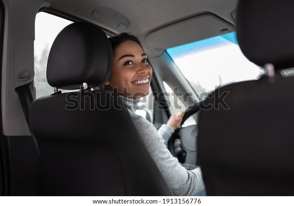 Young woman driving car, trip of business lady in\
sunny weather. Happy cute young african american female holds\
steering wheel, turns in auto and talk with friend or client on\
back seat, empty space