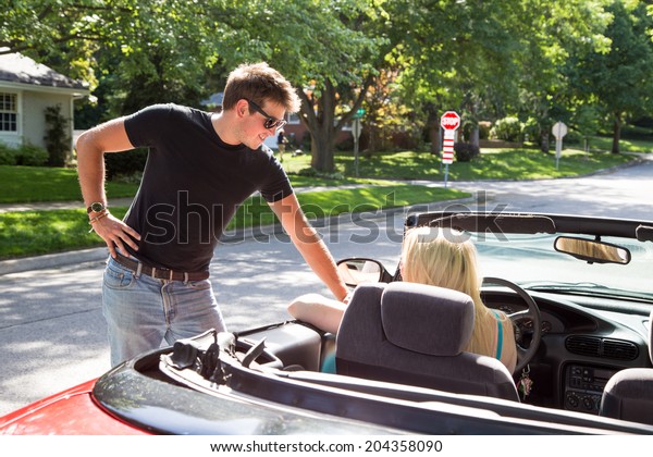 A young woman\
driving a red convertible stops to talk to a young man on the road\
in a residential\
neighborhood.