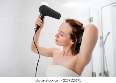 Young woman drying and styling hair with hair dryer, making hairstyle in modern bathroom interior. Beauty routine after morning shower