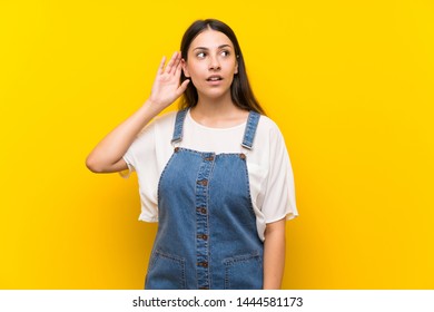 Young woman in dungarees over isolated yellow background listening to something by putting hand on the ear - Shutterstock ID 1444581173