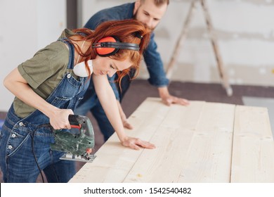 Young Woman In Dungarees Preparing To Use A Power Tool During Renovations To Her Home Watched By A Man In The Background