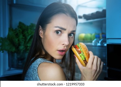 Young Woman Eating Burger From Fridge At Night Kitchen. Diet Fail Concept